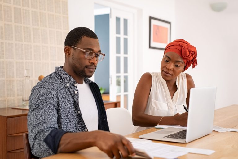 African mature couple managing expenses at home Serious mid adult man and african woman with traditional turban working and writing notes while using laptop. Mature african american couple discussing home finance with bank statement and laptop. Black man and serious woman managing budget and checking accountancy and bills.