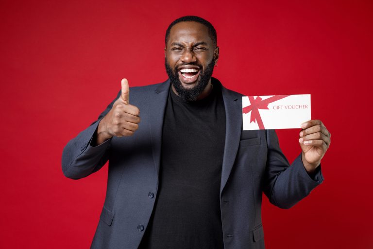 Laughing young african american business man 20s wearing classic jacket suit standing showing thumb up hold gift certificate looking camera isolated on bright red color background studio portrait. Laughing young african american business man 20s wearing classic jacket suit standing showing thumb up hold gift certificate looking camera isolated on bright red color background studio portrait