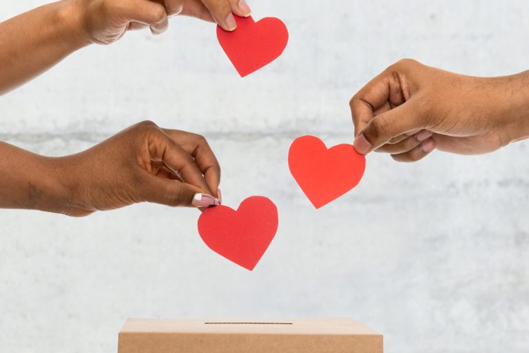 hands putting hearts into donation box on street charity, love and valentine's day concept - close up of hands putting red hearts into donation box over urban steet background